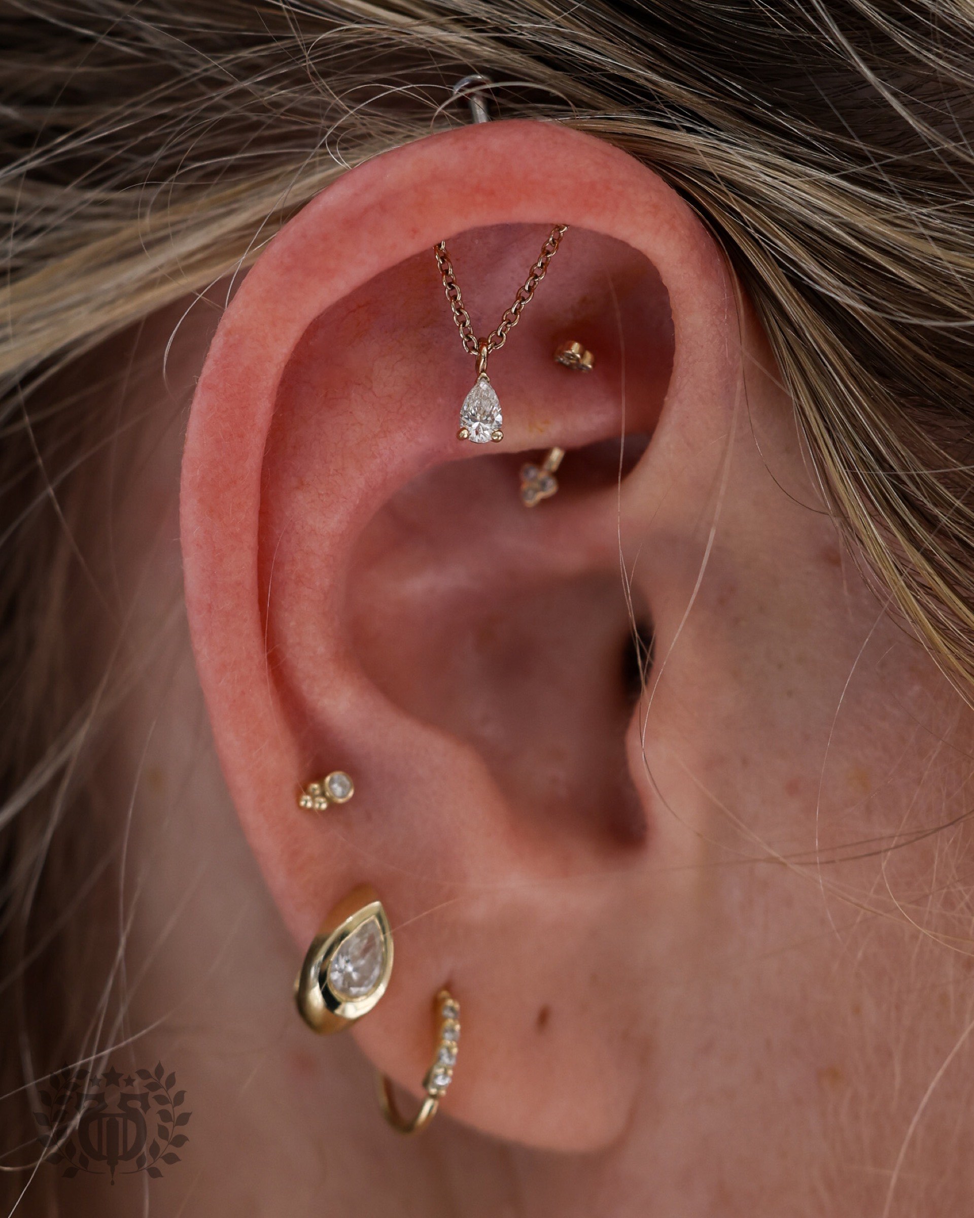 Rook piercing with gold chain and pear-shaped diamond pendant with curated gold lobe jewelry at Apollo Studio