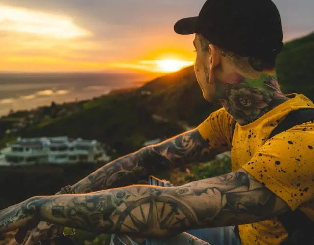 A person with full sleeve tattoo cover ups on both arms and a neck tattoo, gazing at the sunset.